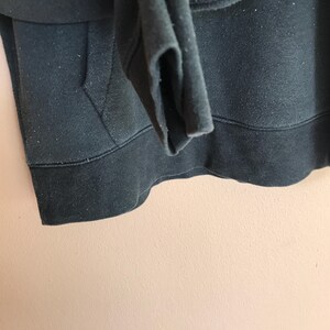 May include: Close-up of a dark green hoodie. The image shows the bottom hem, pocket, and sleeve cuff. The fabric appears to be a soft, slightly worn material. The hoodie has a drawstring and a kangaroo pocket.