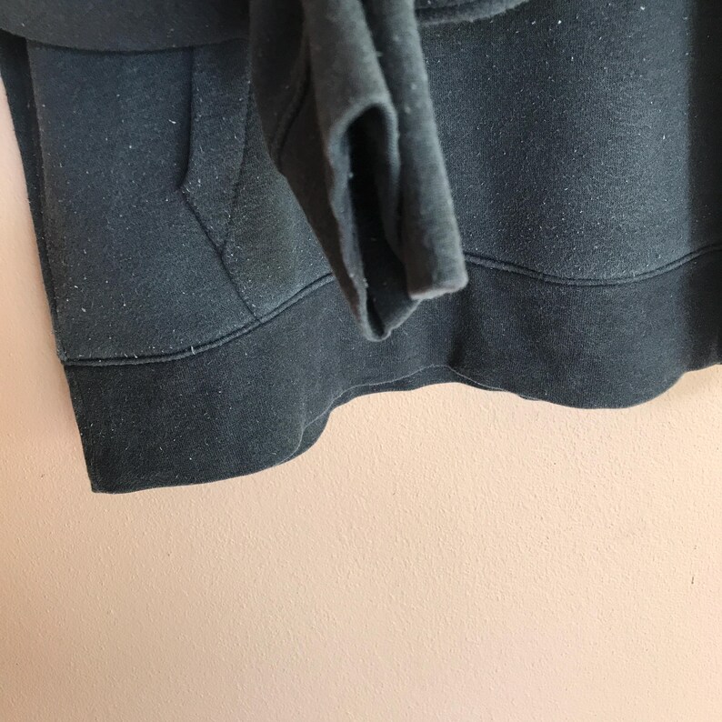 May include: Close-up of a dark green hoodie. The image shows the bottom hem, pocket, and sleeve cuff. The fabric appears to be a soft, slightly worn material. The hoodie has a drawstring and a kangaroo pocket.