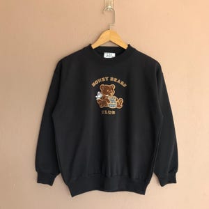 May include: Black sweatshirt with the text "HONEY BEARS CLUB" above a brown teddy bear graphic holding a honey pot. The sweatshirt has a crew neck and long sleeves. The graphic is embroidered in brown and tan.