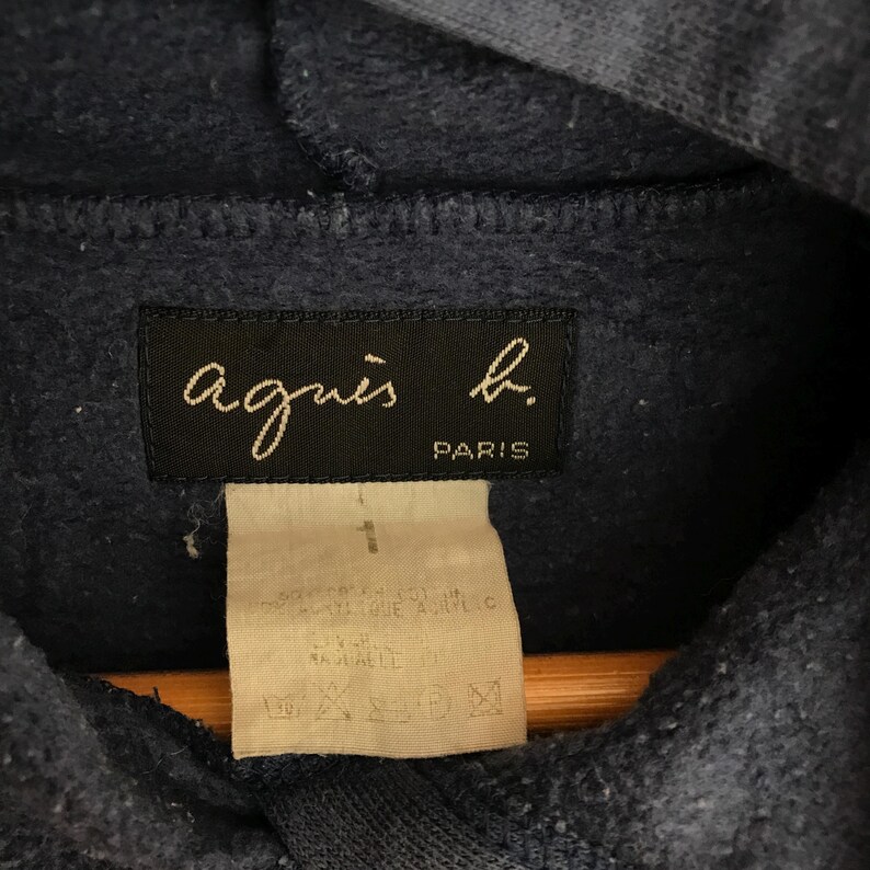 May include: Close-up of a navy blue hoodie with a tag featuring the brand name "agnes b." in cursive and the word "PARIS." A smaller tag below displays care instructions and size information.