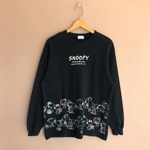Op de afbeelding: Zwarte Snoopy-trui met een ronde hals en lange mouwen. Het woord "SNOOPY" is in het wit op de borst gedrukt, met daaronder "PEANUTS United Feature Syndicate, Inc.". Een rij Snoopy en andere Peanuts-personages is aan de onderkant gedrukt.
