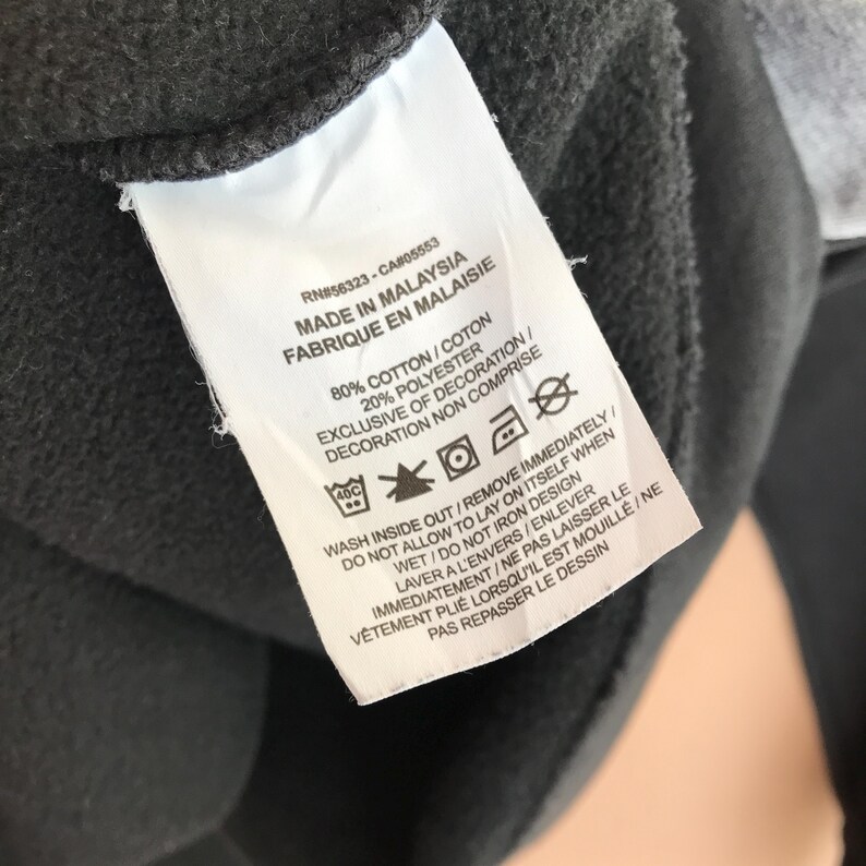 Pu&ograve; includere: Primo piano di un'etichetta bianca con testo nero, attaccata a un capo di abbigliamento grigio scuro. L'etichetta riporta le istruzioni di lavaggio e la composizione del tessuto: 80% cotone, 20% poliestere. C'&egrave; scritto "Made in Malaysia".