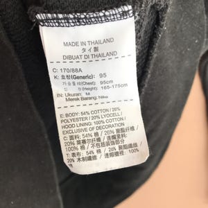May include: A close-up of a black garment tag with text in multiple languages. The tag provides details about the garment's composition, including cotton and polyester percentages, and the brand name Nike. The tag also indicates the garment was made in Thailand.