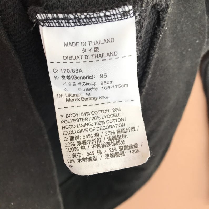 May include: A close-up of a black garment tag with text in multiple languages. The tag provides details about the garment's composition, including cotton and polyester percentages, and the brand name Nike. The tag also indicates the garment was made in Thailand.