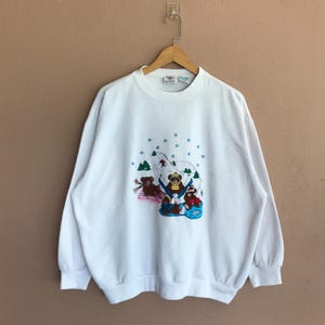 Könnte beinhalten: Weißes Sweatshirt mit Rundhalsausschnitt und langen Ärmeln. Auf der Vorderseite befindet sich ein gesticktes Design von drei Bären, die Ski fahren und rodeln, in einer verschneiten Szene mit grünen Bäumen und blauen Schneeflocken. Das Sweatshirt hängt an einem Holzkleiderbügel.