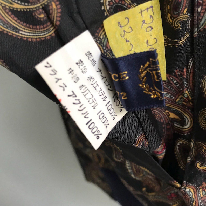 Pu&ograve; includere: Un campione di tessuto paisley nero e marrone con un'etichetta che recita "表地 ナイロン 100% 表地 ポリエステル 100% 中綿 ポリエステル 100% フライス アクリル 100%"