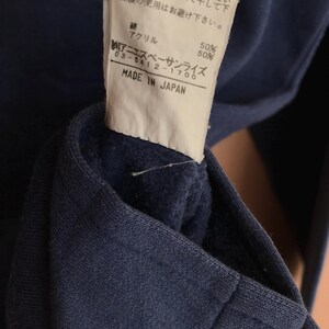 May include: Close-up of a navy blue garment with a tag. The tag has Japanese text, including "MADE IN JAPAN" and the brand name. The fabric composition is listed as 50% cotton and 50% acrylic.