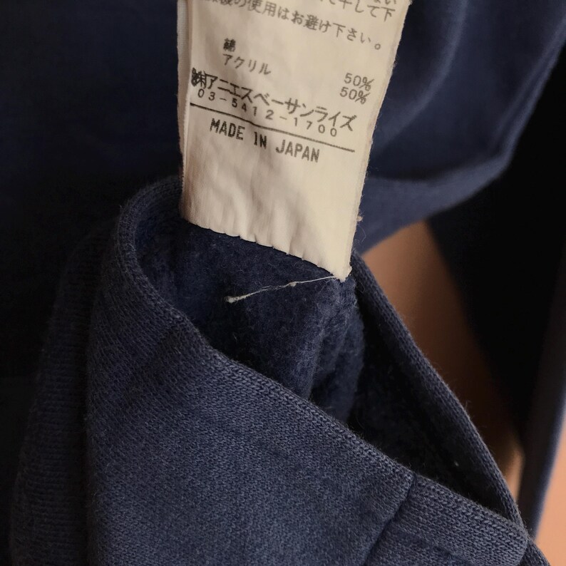 May include: Close-up of a navy blue garment with a tag. The tag has Japanese text, including "MADE IN JAPAN" and the brand name. The fabric composition is listed as 50% cotton and 50% acrylic.