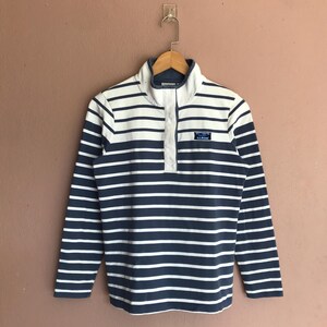 May include: A long-sleeved striped shirt in navy blue and white, hanging on a wooden hanger. The shirt features a button-up collar and a small rectangular patch with text. The shirt is a casual style.