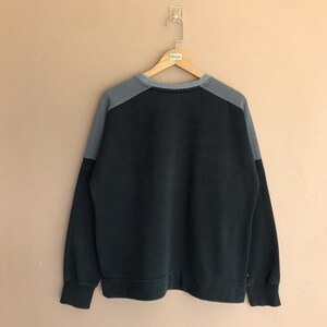 Pu&ograve; includere: Una felpa grigio scuro con pannelli grigi a contrasto sulle spalle, appesa a una gruccia di legno. La felpa girocollo ha maniche lunghe e orlo e polsini a coste. L'indumento &egrave; esposto contro un muro neutro.