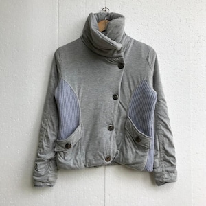 Puede incluir: Una chaqueta acolchada gris con cuello alto y cierre de botones. La chaqueta tiene un detalle de canalé en las mangas y los bolsillos.