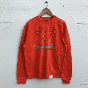May include: An orange sweatshirt with a black diamond graphic and the word "Diamond" in teal script. The words "Supply Co." are below the word "Diamond". The sweatshirt is hanging on a wooden hanger.