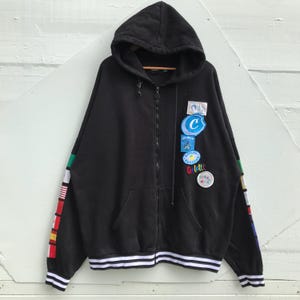 Pode incluir: Casaco de moletom preto com capuz e zíper, com uma faixa de cintura listrada e patches coloridos na manga esquerda. Os patches incluem um círculo azul com um "C" branco dentro, um círculo amarelo com um desenho de sol e um patch azul e branco com a palavra "Gett'it".