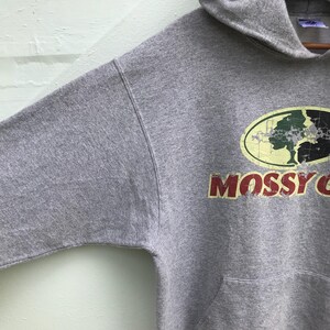 May include: Gray hooded sweatshirt with a distressed Mossy Oak logo featuring a tree and the text "Mossy Oak" in red.