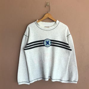 Könnte beinhalten: Ein hellgraues Sweatshirt mit Rundhalsausschnitt und langen Ärmeln. Das Design zeigt zwei schwarze horizontale Streifen über der Brust mit einem kreisförmigen Emblem in der Mitte. Das Emblem hat ein blaues und grünes Design mit dem Buchstaben "X".