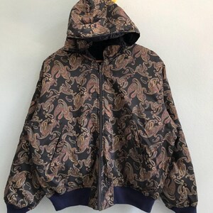 Pu&ograve; includere: Una giacca nera con cappuccio e motivo paisley. La giacca ha una chiusura a zip e una cintura blu scuro.