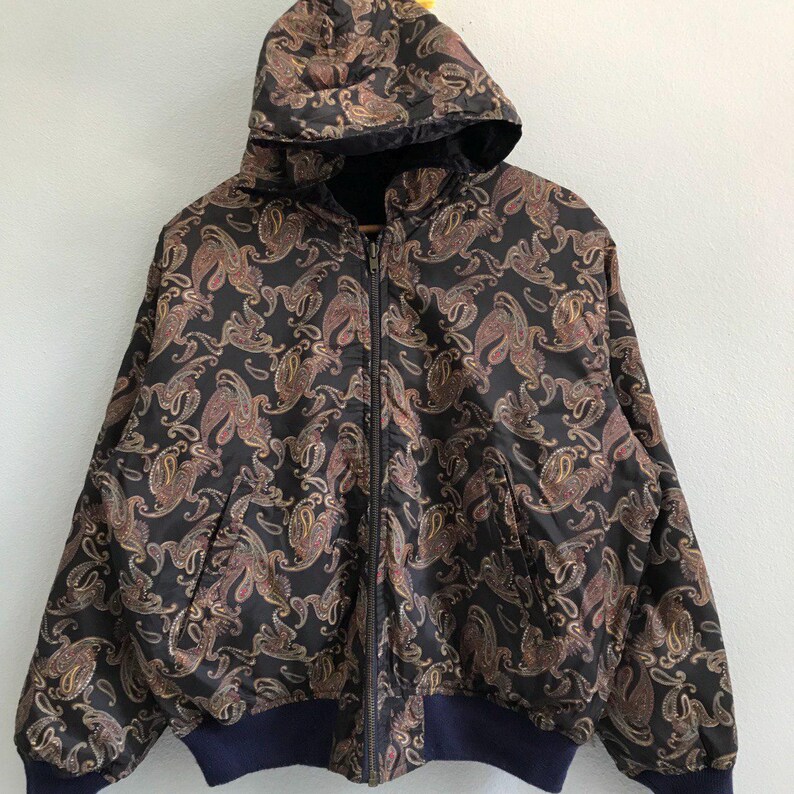 Pu&ograve; includere: Una giacca nera con cappuccio e motivo paisley. La giacca ha una chiusura a zip e una cintura blu scuro.