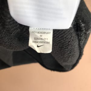 May include: Close-up of a dark grey garment tag with the Nike logo. The tag displays the text "HO180508NAY", "M", "928438-011", and "888408959386". The tag is attached to a dark grey fabric.