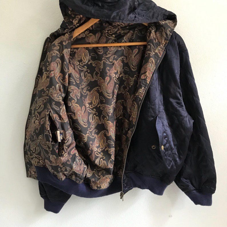 Pu&ograve; includere: Una giacca bomber in satin blu navy con fodera a fantasia paisley. La giacca ha un cappuccio e una chiusura a zip.