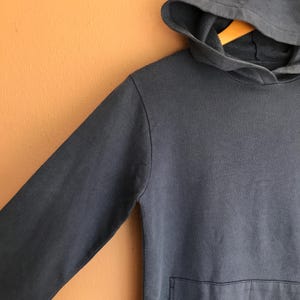 May include: A close-up of a dark blue hoodie with a hood and a front pocket. The hoodie is made of a soft material and is hanging on a wooden hanger against an orange background. The sleeve is visible.