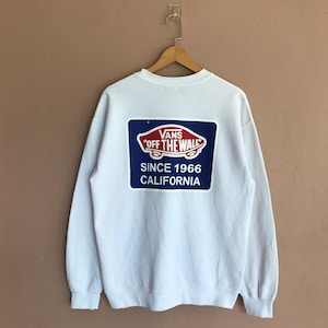 Op de afbeelding: Witte crewneck sweatshirt met een groot blauw en rood Vans logo op de rug. Het logo bevat de tekst "Vans Off The Wall" en "Since 1966 California". De sweatshirt hangt aan een houten hanger.