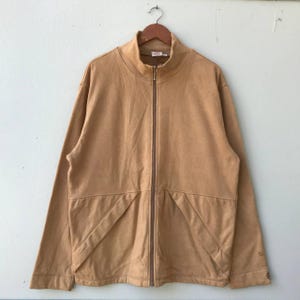 Peut inclure: Veste zippée beige avec un col montant, suspendue à un cintre en bois. La veste a une fermeture éclair sur toute la longueur et deux poches avant. Le tissu semble être un matériau doux semblable à du daim. La veste est de couleur neutre.