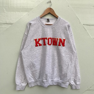 以下が含まれることがあります： 前面に赤い刺繍で「KTOWN」のロゴが入ったグレーのスウェットシャツです。