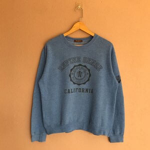May include: A blue sweatshirt featuring the text "Refine Sense" and "California". The design includes a circular emblem with the letters "FA". The sweatshirt has a crew neck and long sleeves.