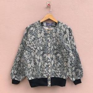 May include: A gray and white leaf-patterned jacket with a black ribbed hem and cuffs. The jacket has a button-up front and is hanging on a wooden hanger against a pink background. The sleeves are long and slightly puffed.