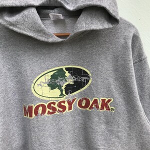 May include: Gray hooded sweatshirt with a distressed Mossy Oak logo featuring a green tree and black background. The logo is centered on the front of the sweatshirt.