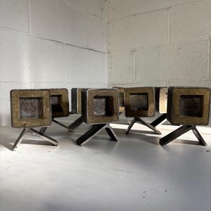 May include: A collection of small, square metal objects with a gold-colored interior and three-legged base. The objects are arranged on a white surface, with a white brick wall in the background. The metal has a slightly aged appearance.