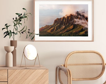 May include: A framed photograph of a mountain range with clouds in the background. The mountain range is a deep brown color and the clouds are white. The photograph is hanging on a light beige wall.