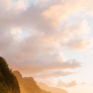 May include: A scenic view of a rocky coastline with lush green vegetation at the base of the cliffs. The sky is a soft, pastel pink and orange with fluffy white clouds. The ocean is calm and blue, reflecting the colors of the sky.