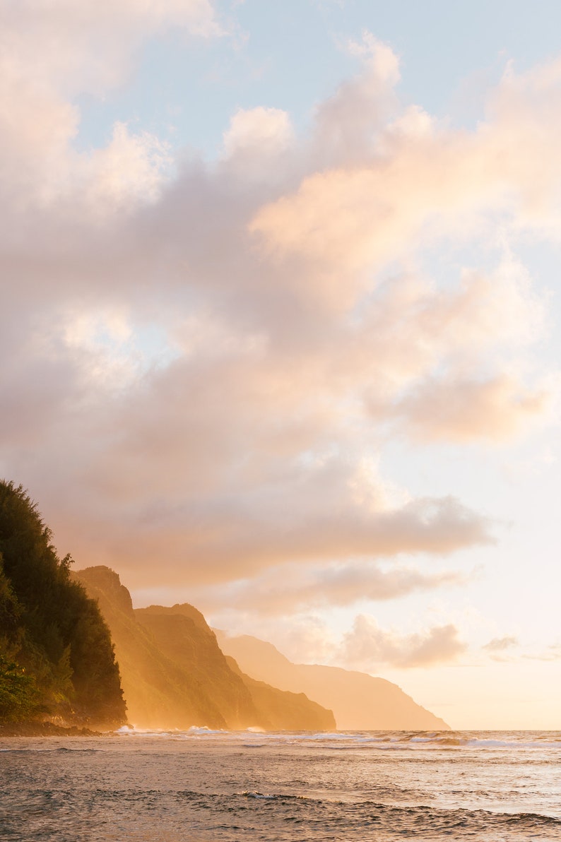 May include: A scenic view of a rocky coastline with lush green vegetation at the base of the cliffs. The sky is a soft, pastel pink and orange with fluffy white clouds. The ocean is calm and blue, reflecting the colors of the sky.