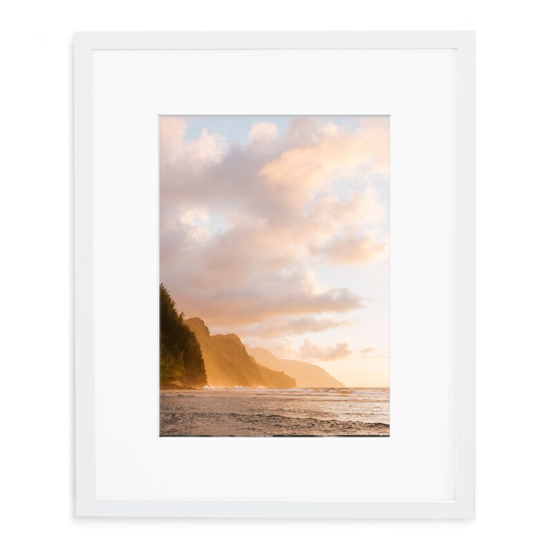 May include: A photograph of a coastline with a lush green mountainside in the background. The sky is a vibrant orange and pink, with fluffy white clouds. The ocean is a deep blue, with waves crashing on the shore.