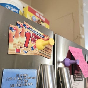May include: A yellow plastic refrigerator magnet shaped like a thumbtack. The magnet is holding up a stack of three pizza coupons that say "1 Free Small Cheese Pizza".