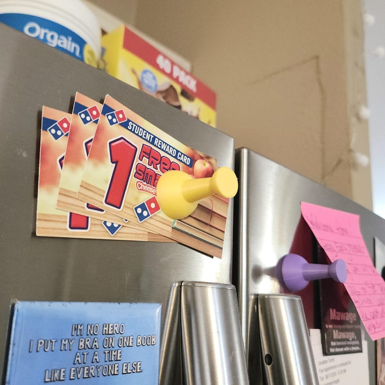 May include: A yellow plastic refrigerator magnet shaped like a thumbtack. The magnet is holding up a stack of three pizza coupons that say "1 Free Small Cheese Pizza".