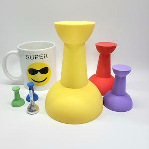 Puede incluir: Un conjunto de cuatro chinchetas de colores en amarillo, rojo, morado y verde. Las chinchetas son de varios tamaños y están dispuestas sobre una superficie blanca. También hay una taza blanca con la palabra "SUPER" y una cara sonriente con gafas de sol en la imagen.
