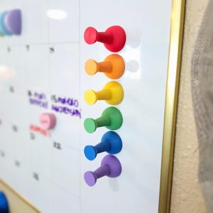 Könnte beinhalten: Ein Satz von sieben bunten Stecknadeln in Regenbogenfarben, die an einer weißen Trockenlöschtafel befestigt sind. Die Stecknadeln sind aus Kunststoff und haben einen abgerundeten Kopf und einen Haken.