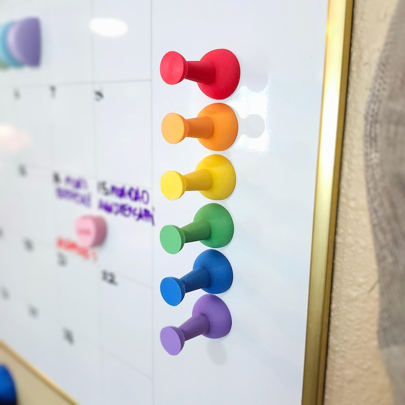 May include: A set of seven colorful push pins in a rainbow pattern, attached to a white dry erase board. The push pins are made of plastic and have a rounded head and a hook.