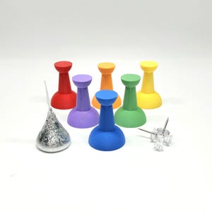 May include: A set of six colorful push pins in red, purple, blue, green, yellow, and orange. The push pins are arranged in a semi-circle on a white surface. There is a silver foil-wrapped chocolate kiss in the center of the image.