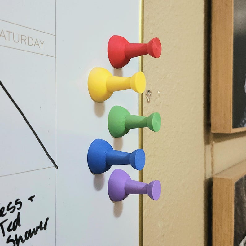 May include: Five colorful push pins in red, yellow, green, blue, and purple are attached to a white board. The push pins are made of plastic and have a rounded head and a small, pointed end.