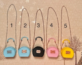May include: Five miniature purses with gold chains. The purses are in different colors: light blue, black, pink, and yellow. Each purse has a small, white label with the number "101" on it.