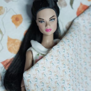 May include: A doll with long black hair is lying on a bed with a white and blue patterned blanket. The doll is wearing a white collared dress with a floral pattern.
