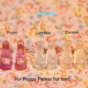 May include: Three pairs of miniature doll shoes in purple, light blue, and caramel colors, labeled as Lollipop for Poppy Parker flat feet. Each pair features a translucent, glossy finish with a textured toe detail and a low-profile flat sole.