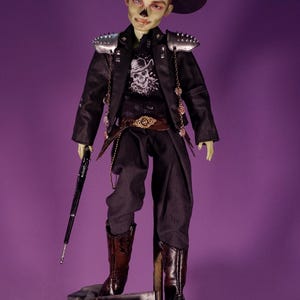 May include: A doll dressed in a black jacket with silver shoulder accents, black pants, and brown boots. The doll wears a black hat and holds a black cane. A skull graphic is on the shirt. The doll stands on a decorative base with a small wooden chest.