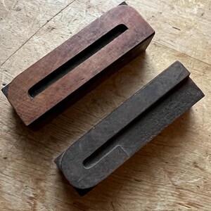 May include: Two vintage wooden printing blocks. One is rectangular with a long, narrow channel. The other is a U-shaped block. Both are dark brown and sit on a wooden surface. These blocks are likely used for letterpress printing.