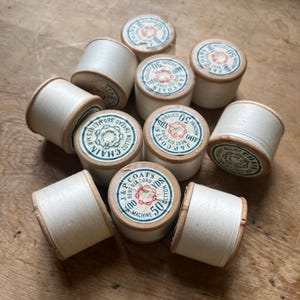May include: A collection of vintage thread spools. The spools are cylindrical, made of wood, and filled with white thread. Each spool has a paper label with text and a red and blue design. The labels indicate the thread type and yardage.