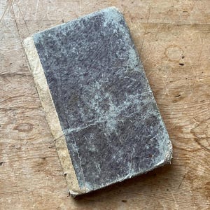 May include: An antique book with a weathered, textured cover in shades of gray and blue-green. The spine is a light beige color. The book is lying on a wooden surface.