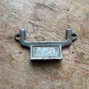May include: A silver metal door handle with a rectangular plate and two small holes on each side. The handle is designed to be used on a cabinet or drawer.
